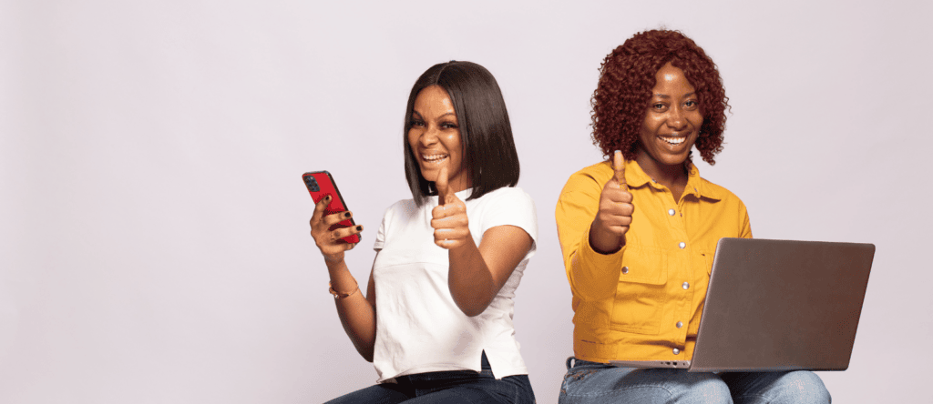Two women-one on a cell phone and one on a laptop-give a thumbs up.