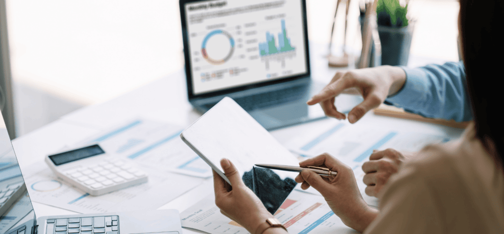 Two people examine a smart tablet. They are above a messy table littered with charts, graphs, and papers. Represents the concept of management consulting SEO.